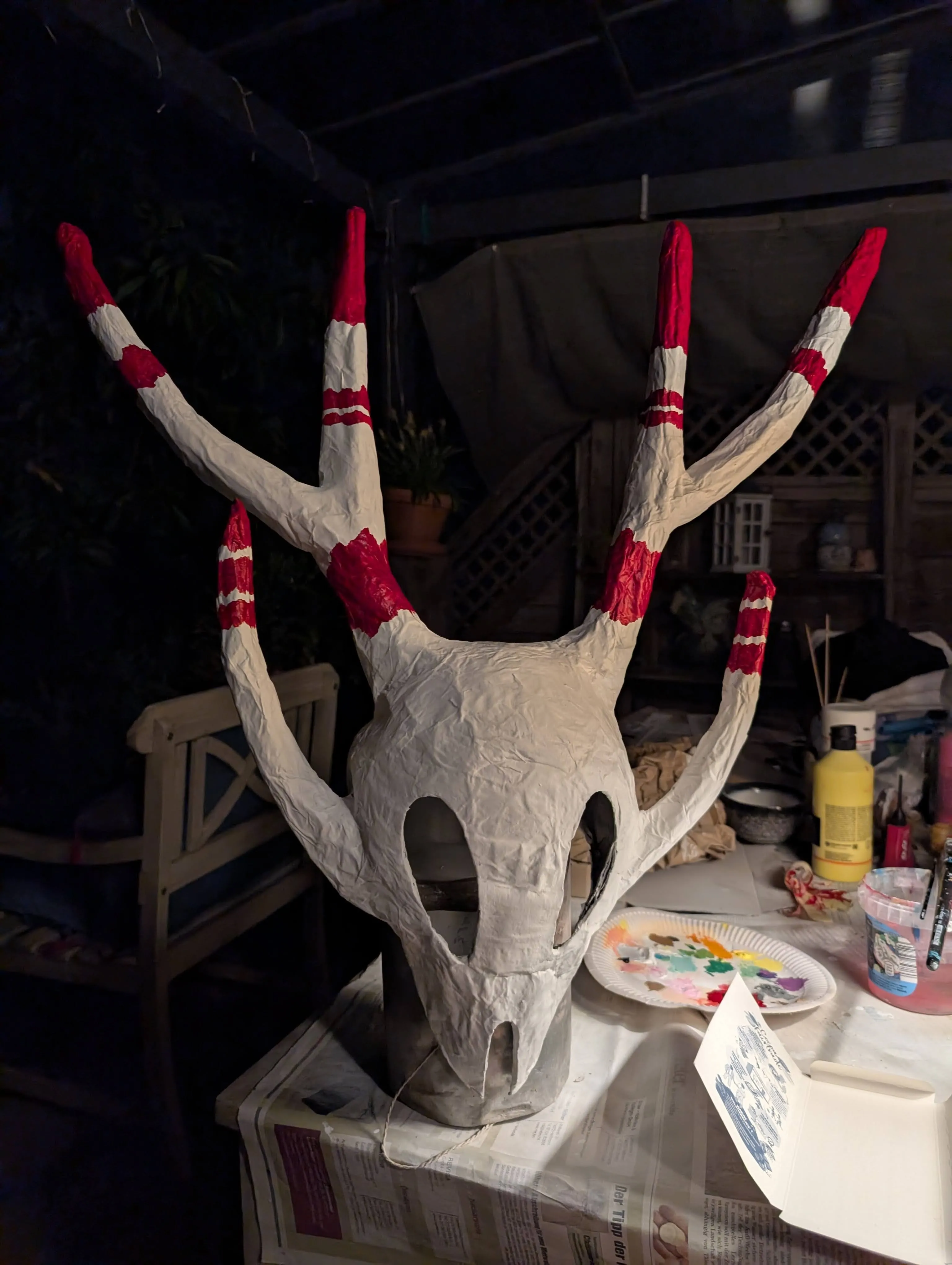 The mask standing on a pot on a messy table being painted in red.